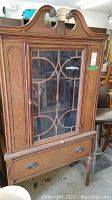 Front view of the tall vintage wooden cabinet showing glass door with wooden geometric overlay and bottom drawer with metal handles.
