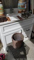 White cabinet with metal crocodile decor on top, small cutting tray, round wicker basket on rectangular woven storage box in front.