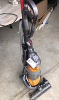 Full view of Dyson DC25 Ball upright vacuum cleaner standing on concrete floor, showing handle, power cord wrapped around, and orange dust container.