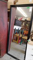 Full view of the tall rectangular mirror with black wood frame showing scratches on frame edges against a background of stacked chairs and furniture.