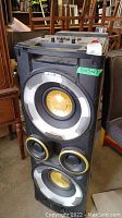 Front view showing the large black stereo unit with multiple speakers with gold cones and a green label with dimensions 15x15x43