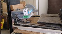 Electric tile cutter with visible King Canada label, motor housing, blade guard, power cord, and cutting platform with slatted surface and white debris tray beneath.