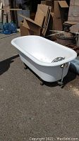 Full view of a white clawfoot tub with metal claw feet on pavement outdoor setting. Cardboard boxes and water jugs in background.
