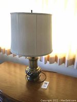 Full view of the brass table lamp with beige shade on wooden surface near window.