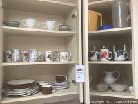 Wide view of cabinet shelves showing assortment of cups, mugs, plates, and servingware including some with floral designs and others with patterned borders.