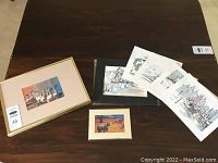 Collection of art prints and sketches shown on a table including framed and loose sheets, featuring various artists.