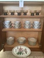 Wide view of dinnerware set showing 12 Noritake coffee cups and saucers, stacks of dessert plates and bowls, 6 glass pedestal dessert dishes, and a large oval serving plate.