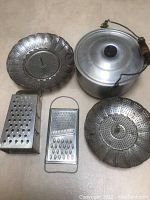 Top view of an aluminum pot with lid and wood handle, two perforated metal steamer inserts, and two different metal graters arranged on a flat surface.