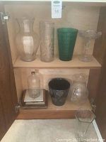 Overview of nine vases including glass, crystal, and ceramic varieties in a wooden cabinet shelf, showing a range of shapes, sizes, and colors.
