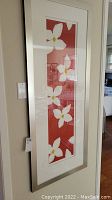Framed art piece with five white flowers vertically arranged on a red background, silver frame and white matting visible. Pieces hanging on wall in room.