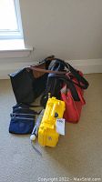 Photo showing the collection of bags, the yellow protective case, and umbrella on carpeted floor near window.