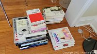 Photo showing multiple stacks of art, photography, fashion, and biography books piled on a wood floor near a white wall and furniture