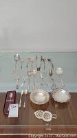 Photo of silver plated serving utensils with various spoons, forks, tongs, and two candy bowls on a reflective glass surface; also includes boxed Oneida cutlery set and two small decorative trivets