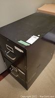 Side view of black 2-drawer metal filing cabinet showing some surface wear and scratches.