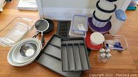 Top-down view of various kitchenware items laid out on wooden floor including glass baking dish, metal bowls, small pan, gray bakeware trays, stacked plastic containers, shakers and utensil holders.