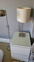 Photo showing white bedside table with two drawers and table lamp with white textured cylindrical shade and metal base.