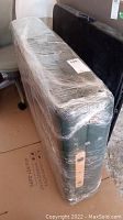 Black folding massage table wrapped in protective plastic, showing side view with wooden handle visible.
