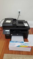 Front view of black HP multifunction printer with paper tray and toner cartridge box placed in front, plugged into wall outlet