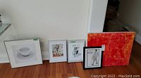 Five pieces of framed art and prints arranged on floor including a photo print of a cup and saucer, three fashion-themed prints, and one large orange-red abstract canvas.
