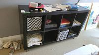 Front view of the black cubed shelf unit showing 8 cubbies with various non-included contents inside (boxes, books, cloth).