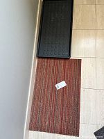 Photo showing black rubber boot tray and reddish striped woven floor mat on tiled floor.