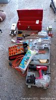 Lot displayed on floor showing all contents: red metal tool box opened with various hand tools, batteries, flashlights, safety gloves, extension cord, and cordless drill in case.