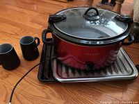 Red Crock-Pot slow cooker with glass lid, placed on metal oven grill pans, two black ceramic mugs beside it on wooden floor.