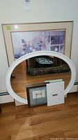 Large framed floral artwork and oval wall mirror along with two smaller framed art pieces leaning against a wall on wooden floor