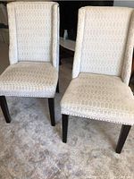 Pair of two upholstered accent chairs with nailhead trim, side by side on carpet showing geometric patterned beige fabric, dark wood legs, and nailhead details.