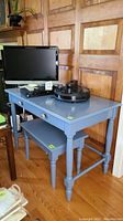 Blue painted desk with flip down keyboard drawer and matching bench under it, situated against wood panel wall and hardwood floor, objects on desk include TV, books, and electronics.
