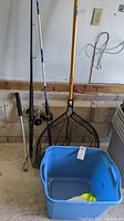 Fishing rods, large fishing net, and blue plastic bin with lures below mounted against a concrete wall in a garage.