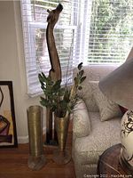 Photo showing the full wooden giraffe sculpture next to two gold metal wire vases, one containing greenery, by a window and couch.