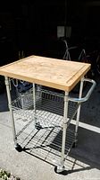 Front and side view of metal kitchen cart showing wooden butcher block top, wire shelves, side hooks, and towel bar.