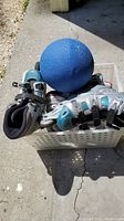 Two pairs of women's rollerblades and a blue rubber bouncing ball resting inside a bin on concrete surface outdoors.