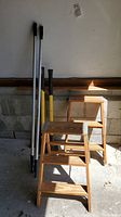Two wooden step ladders, an axe, wood splitter and extendable boat dock hooks in garage setting