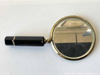 Side view of circular hand mirror with black hinged handle and gold-tone frame on reflective surface.