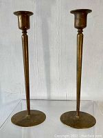 Front view of pair of tall bronze candlestick holders on clear surface, against plain light wall background