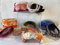 Multiple vintage and antique hats and fascinators including floral fabric bucket hat, red velvet hat with pink feathers, black hat with pink loop decoration, and several veiled hats in burgundy, blue, and other colors.