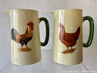 Front view of both cream-colored ceramic pitchers with green handles, one featuring a rooster on the body and the other a hen.