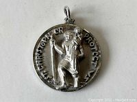 Front view of the silver medallion showing a raised figure of St Christopher carrying a child, with surrounding text.