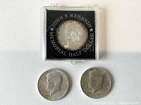 Three Kennedy Half Dollar coins, one in protective case showing front side with JFK profile and inscription.
