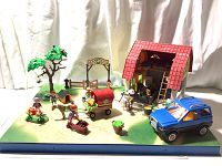 Diorama playset showing assembled Playmobil country scene with stable, vehicle, wagon, figures, tree, and accessories on green base inside display case.