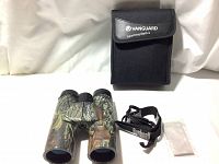 Binoculars, black carrying case, neck strap, and white cleaning cloth displayed on white surface against white backdrop.