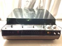 Front view of the audio unit with closed dust cover showing the woodgrain base, chrome trim, AM/FM radio tuner scale, and controls.