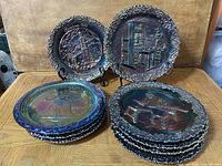 Ten vintage Fenton carnival glass plates stacked and displayed, showing the iridescent surface and embossed scenes including historical and thematic motifs.
