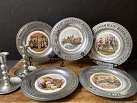 Six commemorative plates The Great American Revolution 1776 with detailed colored historical scenes displayed on plate holders and lying flat on a table.