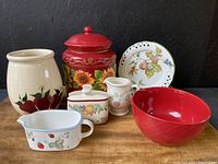 Full lot showing 8 ceramic kitchenware items on a wooden surface against a dark background. Items include cookie jar, bowl, plate, napkin holder, creamers, and vases with fruit and floral motifs.