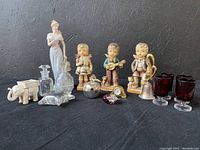 Photo showing all items in the lot arranged in a row, including the figurines, glass bottles, glasses, elephant votive holder, and various collectible items