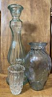 Three clear glass items on a wooden surface including a tall candle holder, a rounded vase, and a small textured candle holder.