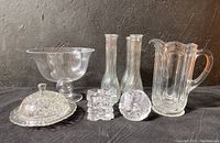Photo shows all items in the lot arranged together on black surface with dark background: footed bowl, pitcher, four slender bud vases, covered dish, two votive candle holders; includes visible flaw on pitcher handle.